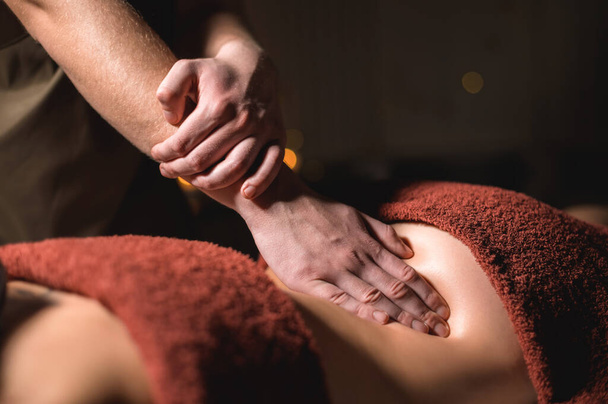 Close-up male hand of a professional physiotherapist masseur doing lumbar massage to a female patient in a massage luxury room with dim light - Foto, imagen