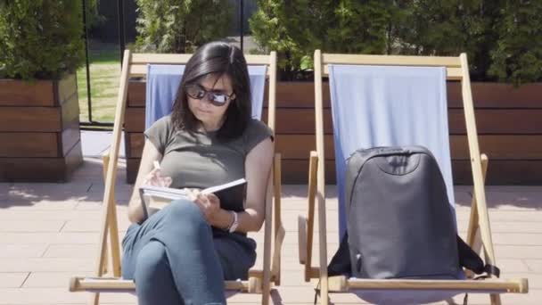 Young woman in sunglasses taking notes in notebook outdoors - Filmmaterial, Video