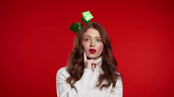 Thinking woman in Christmas head wreath looking up and around on red background. Worried contemplative face expressions. Beautiful young model with beautiful hairstyle. - Footage, Video