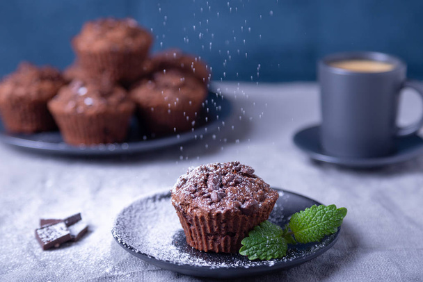 Chocolate muffins with mint on a black plate, strewed with icing sugar. Homemade baking. In the background is a cup of coffee and a plate with muffins. Blue background. Selective focus, close up. - Фото, изображение