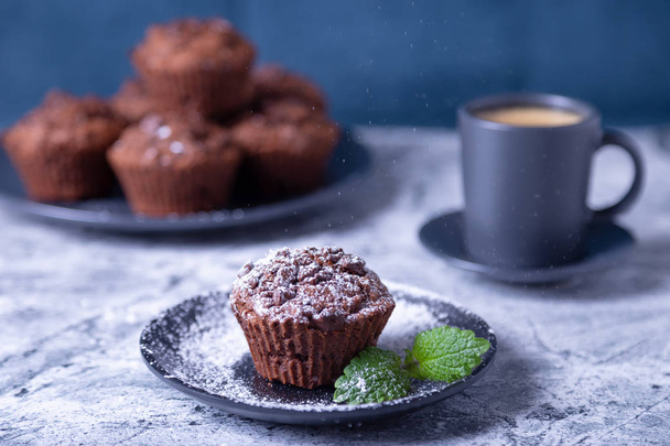 Chocolate muffins with mint on a black plate, strewed with icing sugar. Homemade baking. In the background is a cup of coffee and a plate with muffins. Marble table and blue background. Selective focus, close up. - 写真・画像