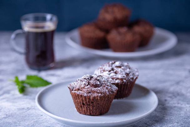 Chocolate muffins with mint on a black plate, strewed with icing sugar. Homemade baking. In the background is a cup of coffee and a plate with muffins. Marble table and blue background. Selective focus, close up. - Foto, Imagen