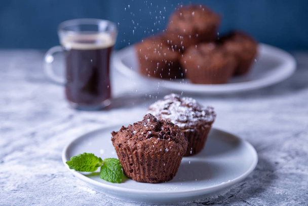 Chocolate muffins with mint on a black plate, strewed with icing sugar. Homemade baking. In the background is a cup of coffee and a plate with muffins. Marble table and blue background. Selective focus, close up. - Фото, изображение