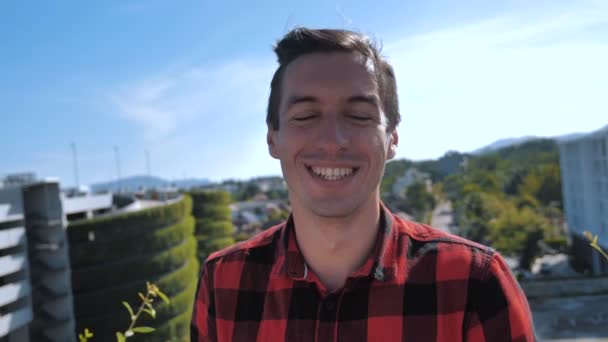 Close-up Portret van vrolijke gelukkige jonge man in Plaid Shirt lachen kijkend naar camera buiten op het dak op Urban City achtergrond - Video