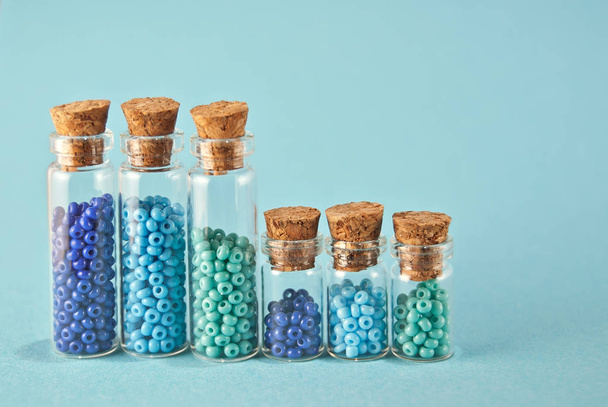 Purple and cyan beads in glass jars on bright blue background. Beads in a transparent container with a wooden cork. The concept of orderliness, balance and chaos. - Zdjęcie, obraz
