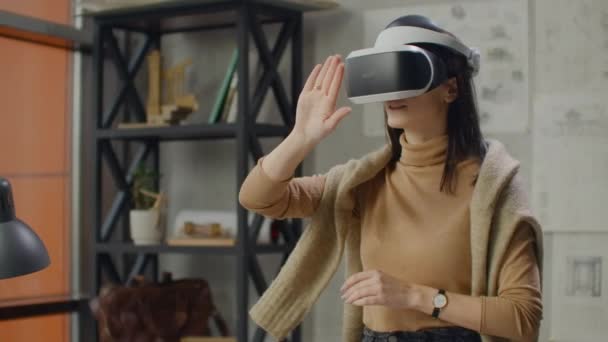 Mujer joven diseñadora en oficina en casco de realidad virtual con manos realiza movimientos imitando el trabajo de interfaz gráfica
 - Imágenes, Vídeo