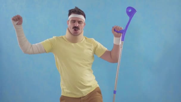 Funny mustachioed man after a sports injury with crutches dancing in front of the camera standing on a blue background - Πλάνα, βίντεο