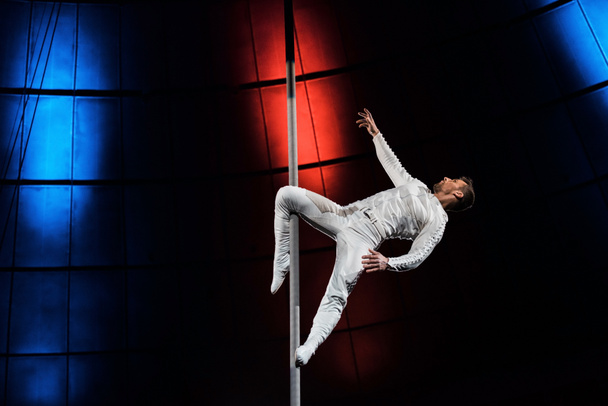 visão lateral de acrobata forte segurando poste metálico com perna enquanto se apresenta no circo
  - Foto, Imagem