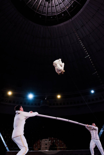 acróbatas sosteniendo poste y mirando a gimnasta saltando en circo
 - Foto, imagen