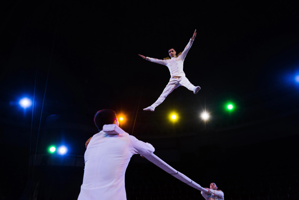 low angle view of handsome air acrobat with outstretched hands jumping near pole in arena of circus  - Photo, Image