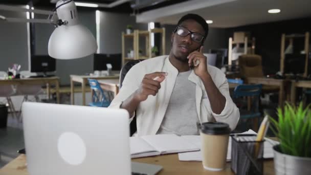 Afrikalı adam cep telefonuyla konuşuyor ve modern ofiste kalemle oynuyor. - Video, Çekim