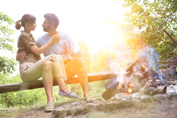 Attractive couple sitting on bench in front of camp fire showing affection with yellow lens flare   - Foto, afbeelding