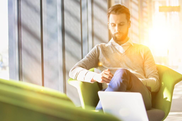 Young attractive businessman using smartphone while sitting and waiting in office lobby with yellow lens flare in background - Photo, Image