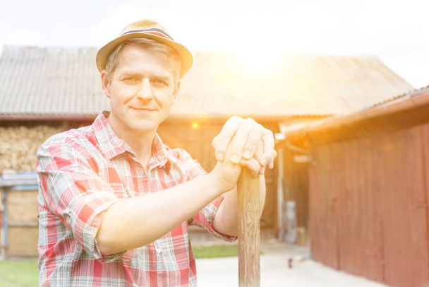 Mature farmer holding shovel in barn with yellow lens flare in background  - Фото, изображение
