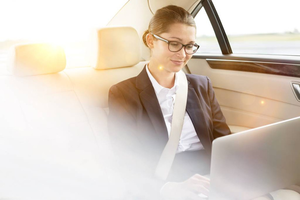 Young attractive businesswoman working on her laptop while sitting inside the car with yellow lens flare in background  - Zdjęcie, obraz