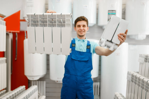 Instalatér v uniformě ukazuje ohřívač vody v instalatérském skladu. Muž kupující sanitární inženýrství v obchodě - Fotografie, Obrázek