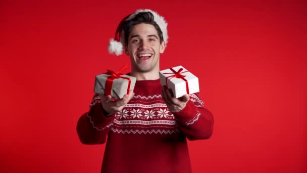 Young handsome man smiling and holding gift box on red studio background. Guy in Santa hat and ugly christmas sweater. New year mood. - Video