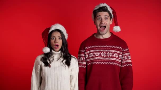 Amazed european man and woman shocked, saying WOW. Young couple in Santa hats surprised to camera over red background. - Materiał filmowy, wideo