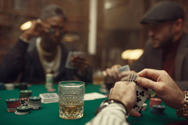Δύο παίκτες πόκερ με κάρτες που κάθονται στο τραπέζι τυχερών παιχνιδιών με πράσινο πανί στο καζίνο. Παιχνίδια εθισμού στην τύχη, κινδύνου, τυχερών παιχνιδιών. Ανδρικές λιχουδιές με ουίσκι και πούρα - Φωτογραφία, εικόνα