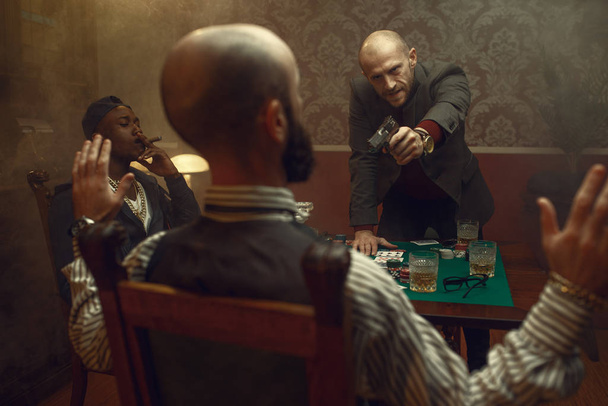 Poker player with gun wants to take the winnings, showdown with opponents in casino, risk. Games of chance addiction. Men with whiskey and cigars in gambling house - Photo, Image