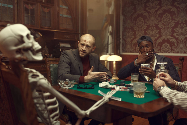 Jogadores de poker masculinos e esqueleto na mesa de jogos com pano verde, diversão, cassino. Jogos de vício casual, risco, casa de jogo. Homens lazeres com uísque e charutos
 - Foto, Imagem
