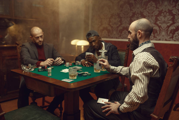 Male player cheating in poker at gaming table, casino. Games of chance addiction, risk, gambling house. Men leisures with whiskey and cigars - Photo, Image