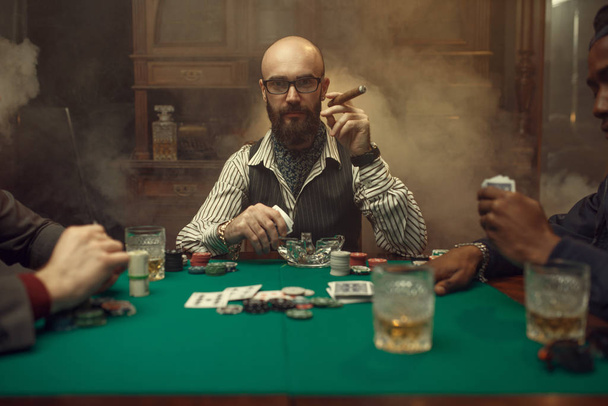 Γενειοφόρος παίκτης πόκερ με πούρο, καζίνο. Παιχνίδια εθισμού στην τύχη. Ο άνθρωπος δελεάζεται από τζόγο. Κάρτες, μάρκες και ουίσκι στο τραπέζι με πράσινο πανί - Φωτογραφία, εικόνα