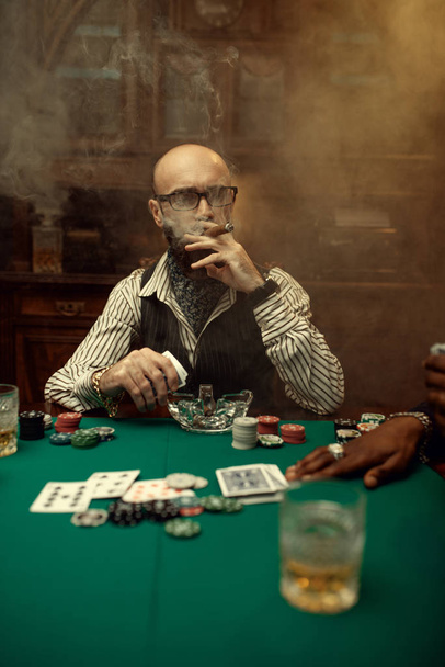 Jugador de poker barbudo con cigarro, casino. Juegos de adicción al azar. El hombre descansa en la casa de apuestas. Tarjetas, fichas y whisky en la mesa de juego con paño verde
 - Foto, Imagen