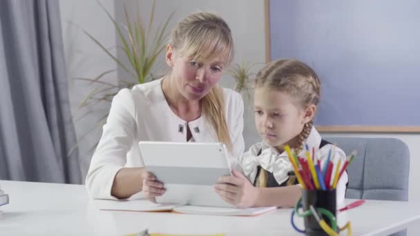 Portrait of middle aged Caucasian woman and little schoolgirl sitting at the table with tablet. Tutor teaching schoolgirl at home. Education, private teaching. Camera moving from left to right. - Materiaali, video