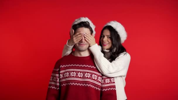 Woman closes eyes of her beloved boyfriend before surprise him. Couple in Santa hat on red studio background. Love, holiday, happiness concept. - Кадры, видео
