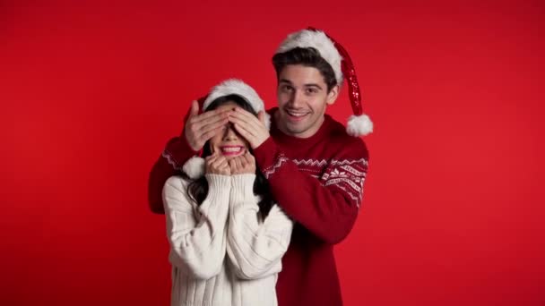 Man closes eyes of his beloved girlfriend before surprise her. Couple in Santa hat on red studio background. Love, holiday, happiness concept. - Кадры, видео