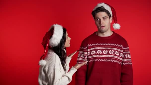 Young woman in Christmas hats emotionally scolds her boyfriend on red background in studio. Bored man rolling his eyes. Concept of conflict, problems in relationships. - Séquence, vidéo