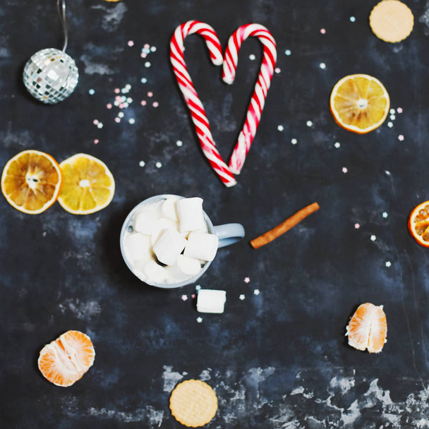 Christmas composition. A cozy mug with marshmallows, candy canes, oranges and decorations on black background. top view - Foto, Bild