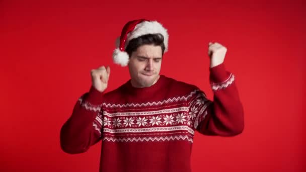 Hezký evropský muž v Santa klobouku a ošklivý svetr s úsměvem a tancem ve vynikající náladě na červeném pozadí. Vánoce, Silvestr, párty koncept. - Záběry, video