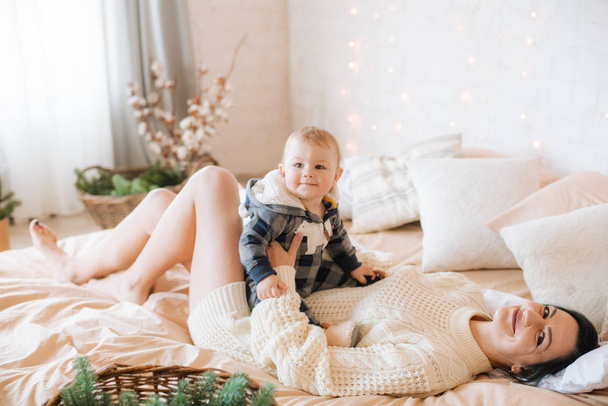Young mother and little toddler son in a Scandinavian style bedroom decorated with Christmas garlands and needles on a large bright bed. Christmas mood - Foto, Imagem