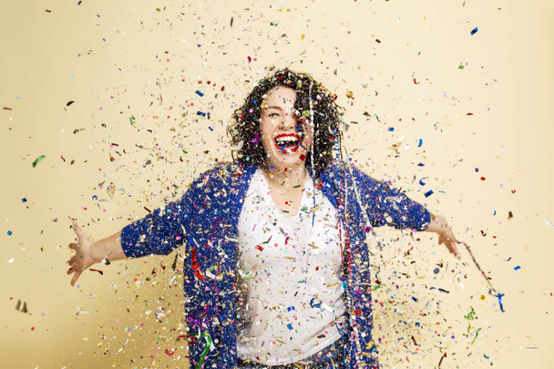 Young beautiful woman with curly hair sprinkled with confetti laughs. Yellow background. - Fotografie, Obrázek