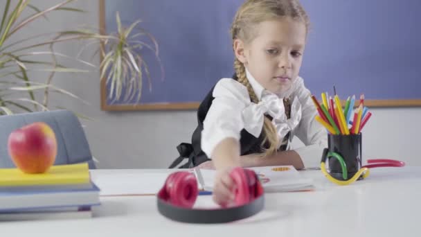 Portrait of pretty Caucasian girl putting on earphones, making victory gesture and shaking head. Happy cheerful child resting as doing homework at home. education, happiness, studying. - Záběry, video