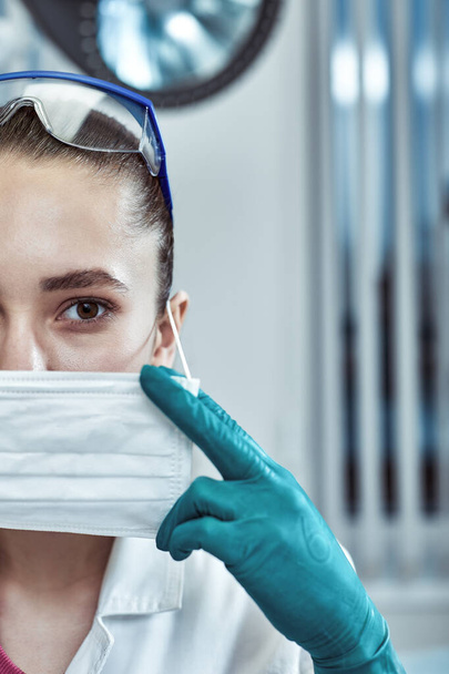 fiatal gyönyörű nő orvos fehér egyenruhában és maszk az arcán közelről klinikai háttér - Fotó, kép