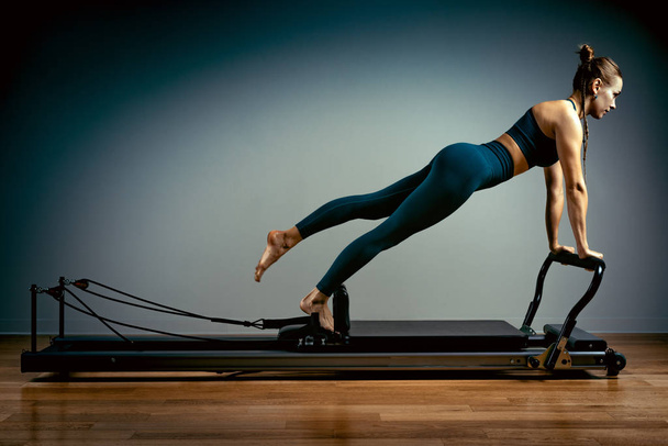 Jong meisje doet pilates oefeningen met een reformer bed. Mooie slanke fitnesstrainer op reformer grijze achtergrond, low-key, art light. Fitness concept - Foto, afbeelding