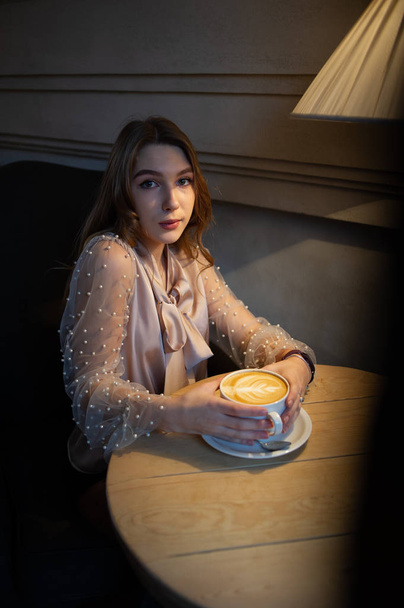 Una joven y bonita mujer en la cafetería. Grunge. Chica en un café con una blusa beige. Una chica bebe café en un café. Latte, Cappuccino
 - Foto, Imagen