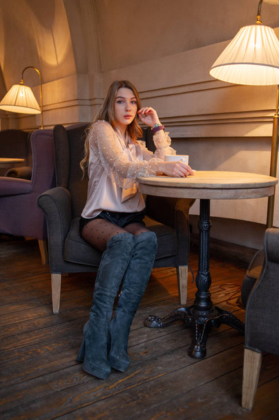 Nuori kaunis nainen kahvilassa. Grunge! Tyttö kahvilassa beigessä puserossa. Tyttö juo kahvia kahvilassa. Latte, Cappuccino
 - Valokuva, kuva