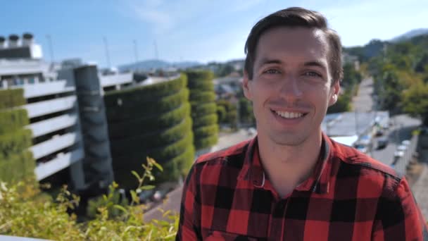 Close-Up Portrait Of Joyful Young Man in Plaid Shirt Smiling Laughing Looking at Camera Outdoors on the Roof at Urban City Background - Footage, Video