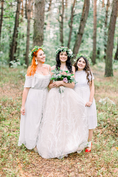 Pretty bride and two bridesmaids in white dresses smiling and looking at camera while standing on the background of pine forest. Stylish and rustic wedding. Marriage concept - Fotografie, Obrázek