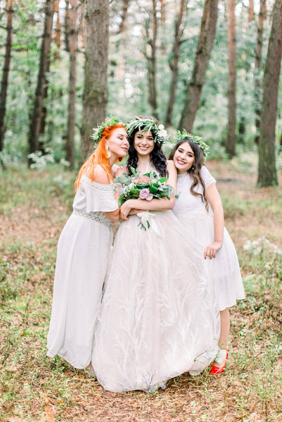 Pretty bride and two bridesmaids in white dresses smiling and looking at camera while standing on the background of pine forest. Stylish and rustic wedding. Marriage concept - Fotoğraf, Görsel
