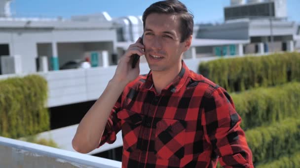 Jeune homme souriant parlant téléphone portable sur le toit. Portrait d'un homme d'affaires ambitieux parlant téléphone portable à l'extérieur à l'arrière-plan urbain. Entrepreneur discuter d'affaires au téléphone à l'extérieur
. - Séquence, vidéo