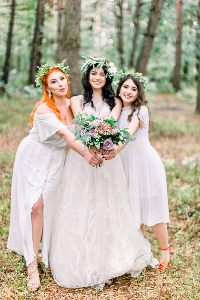 Beautiful bride in stylish rustic dress and wreath is holding flower bouquet in her hands. Two pretty bridesmaids in white dresses and wreaths are hugging the bride. Rustic wedding in the forest - Fotoğraf, Görsel