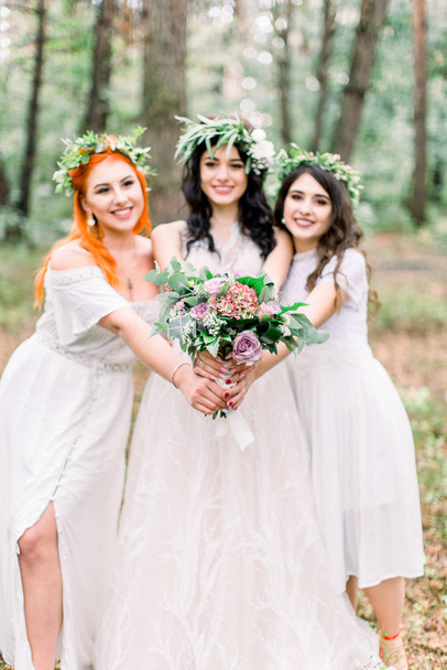 Beautiful bride in stylish rustic dress and wreath is holding flower bouquet in her hands. Two pretty bridesmaids in white dresses and wreaths are hugging the bride. Rustic wedding in the forest - Photo, image