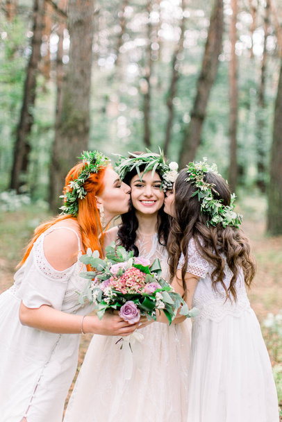Two beautiful bridesmaids kiss brides cheeks while they stand in the forest. Girls in floral wreaths and rustic dresses posing in the forest - Photo, image