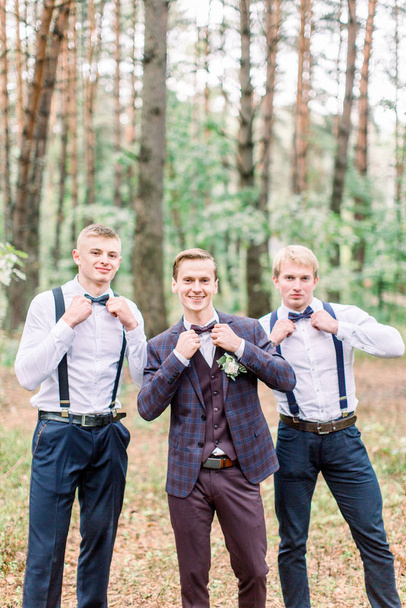 Wedding portrait of groomsmen and handsome groom, touching bow ties. Young cheerful friends groom and groomsmen outdoors in the forest - Valokuva, kuva