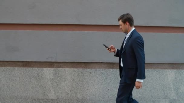 Hombre de negocios con vista lateral caminando con teléfono. Hombre usando el teléfono cerca de edificio moderno - Metraje, vídeo